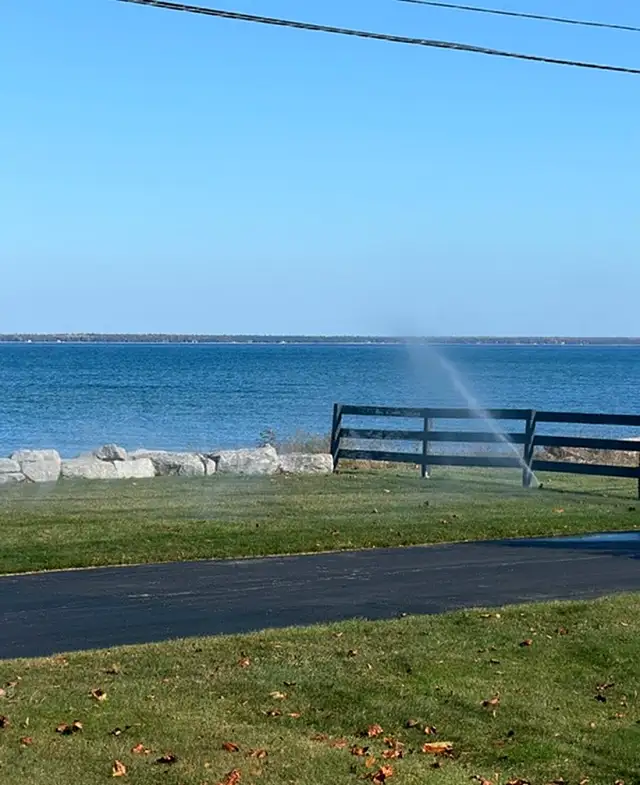 A sprinkler blowing out beside a wooden fence on a lawn along the bay