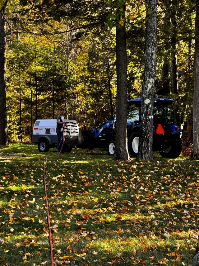 The trailer air compressor and a blue tractor set up under the trees for an irrigation blowout