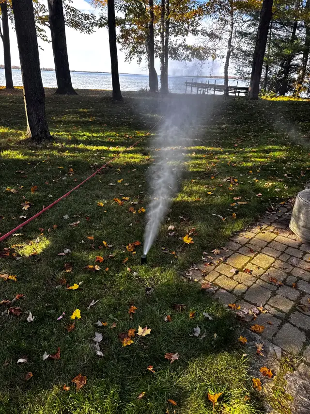 A sprinkler head blowing out a plume of air and mist on a lakefront lawn beside a brick path