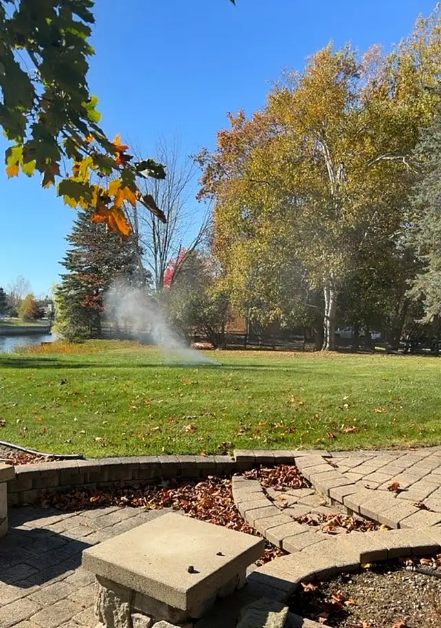 A sprinkler blowing out on a lawn behind a paver patio under autumn trees