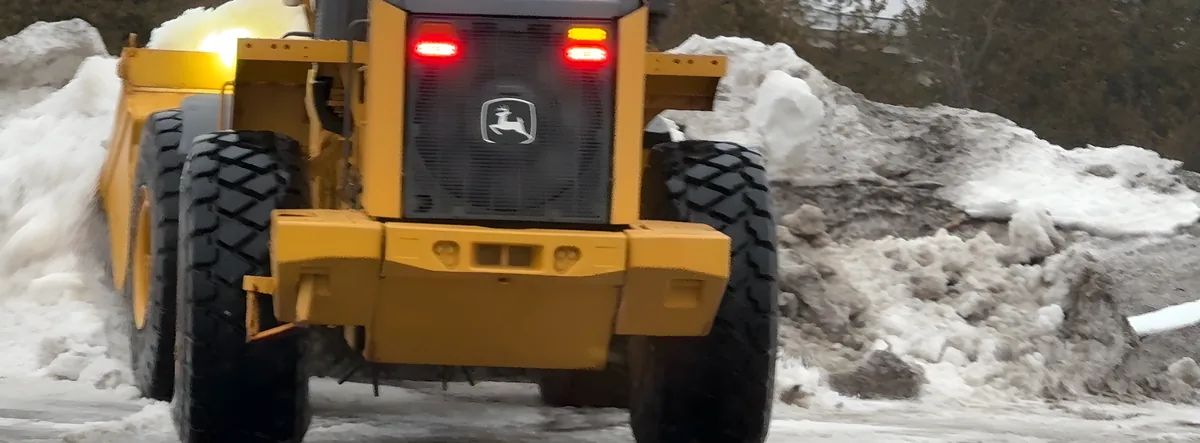 Rear view of a yellow wheel loader with its work lights on beside snow piles
